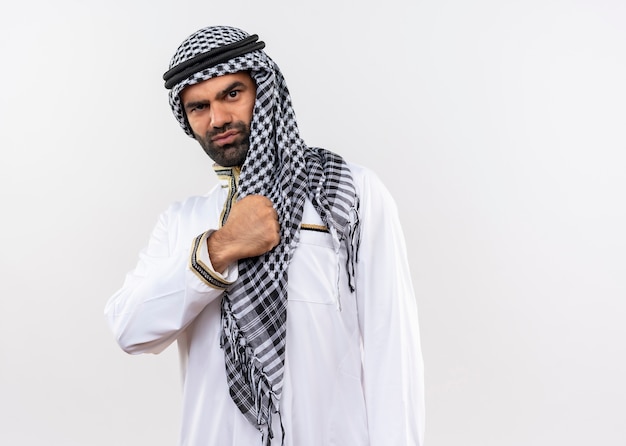 그의 가슴 위에 주먹으로 전통적인 착용을 입은 아랍 남자는 흰 벽 위에 자기 만족과 자신감을 가지고 서 있습니다.