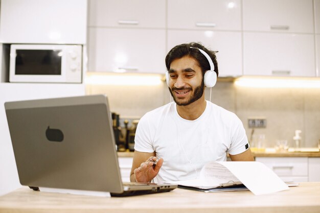 Arabic man watching online webinar, sitting in a kitchen with computer, enjoying distance learning.