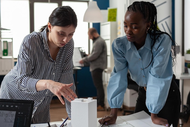 Architect colleagues discussing design of white foam building model in modern architectural office. Team of two professional architects working on blueprints on desk.