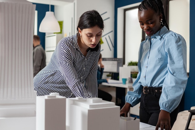 Architectural engineers working together reading construction plans standing next to table with foam model maquette. Team of two architects doing teamwork looking down at blueprins.