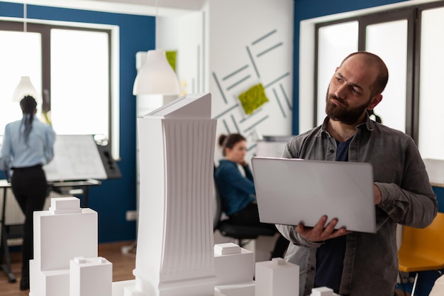 Architectural inspector holding laptop tilting head looking at maquette of skyscraper in residential project. Architect thinking about design improvements to urban planning scale model.