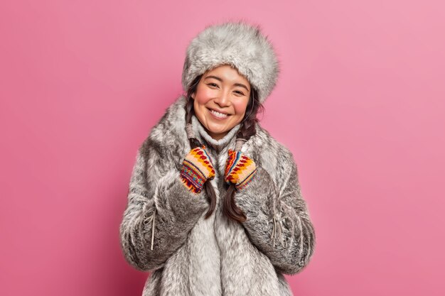 冬服の笑顔の北極圏の女性は、寒い気候の笑顔に広く住んでいますピンクの壁に隔離されたおさげ髪を優しく保持します