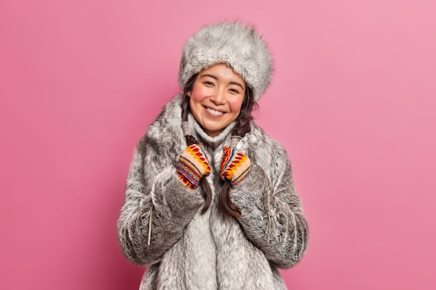 Arctic woman in winter clothes smiles broadly lives in cold climate smiles gently holds pigtails isolated over pink wall