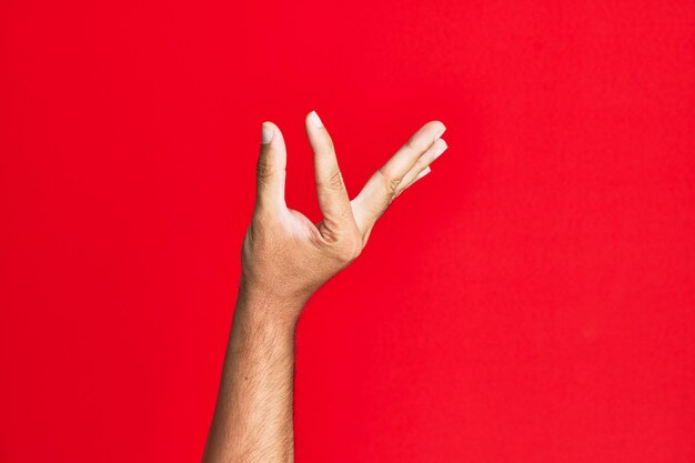 赤い孤立した背景の上に白人の白人青年の腕が、空間を示す指でオブジェクトを保持している目に見えないものを選んで取る