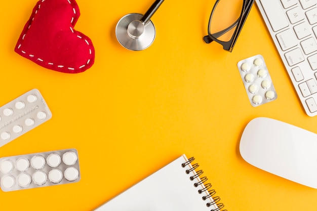 Arrangement of blister packed medicines; stitched heart shape; spiral notepad; wireless keyboard; mouse; spectacles; stethoscope over yellow backdrop