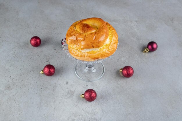 Arrangement of christmas tree toys around a glass pedestal with a sweet bun on marble table.