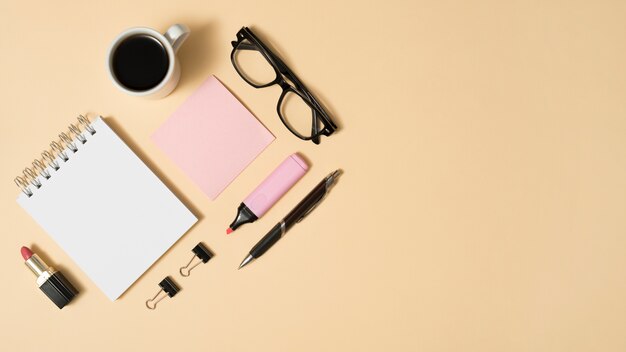 Arrangement of coffee cup; eyeglass; lipstick; with office stuff on beige backdrop