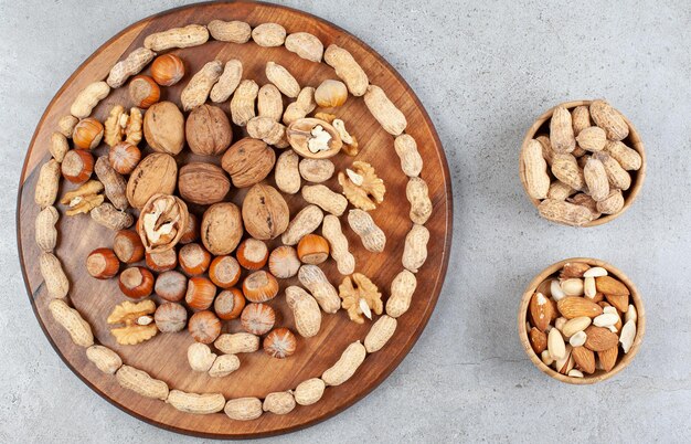 An arrangement of various nut types on wooden board with bowls of peanuts, almonds and pistachios on marble surface.