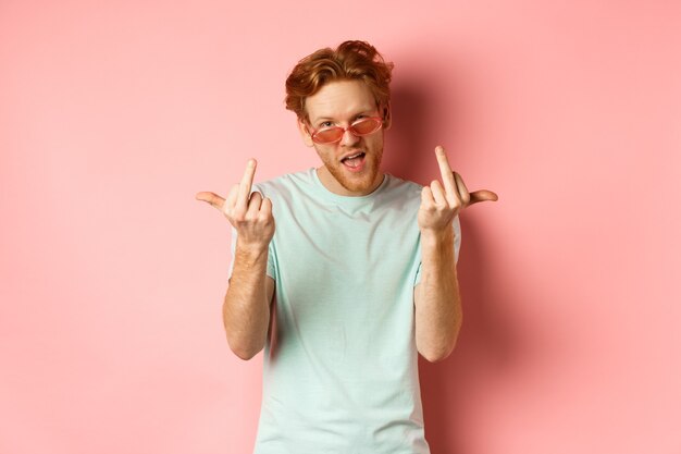 Arrogant and careless redhead man in sunglasses dont give a fuck showing middle fingers at camera an...