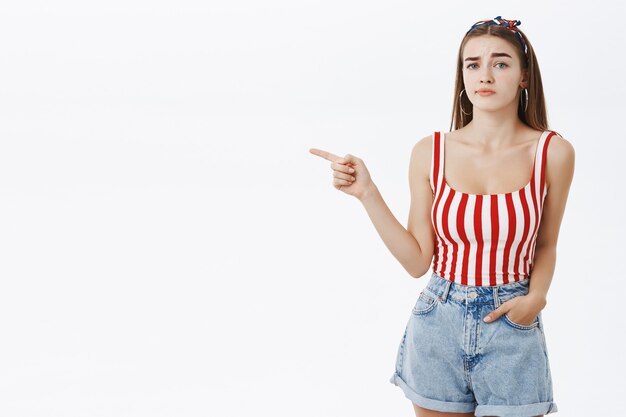 Arrogant snobbish hot woman in pin-up summer outfit raising one eyebrow in disbelief pointing left holding hand in pocket and gazing with indifference, being unimpressed and doubtful