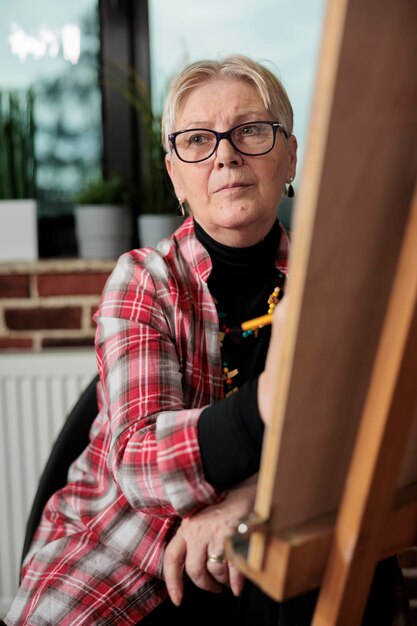 Free photo art classes for seniors. pensive mature woman sitting in front of easel looking aside, listening to teacher during drawing lesson, learning sketching in small groups. older people and creativity