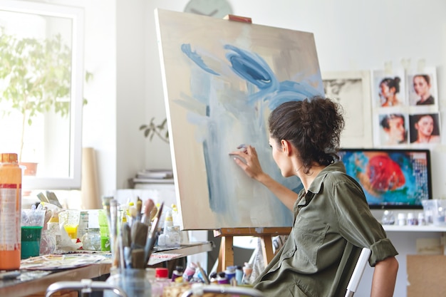 Концепция искусства, творчества, хобби, работы и творчества. Вид сзади занятой художницы, сидящей на стуле перед мольбертом, рисующей пальцами, используя бело-синюю масляную или акриловую краску