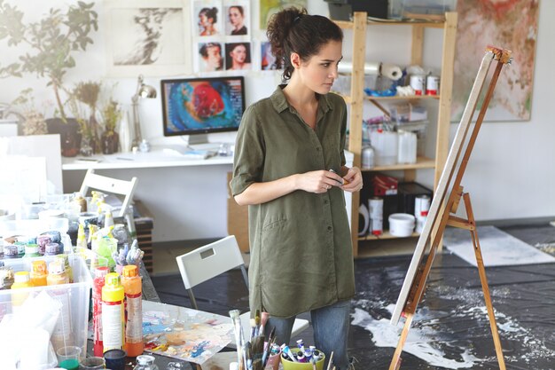 Art and inspiration. Indoor shot of hesitant young female artist wearing jeans and khaki shirt standing in spacious workshop interior in front of easel, evaluating painting that she just finished