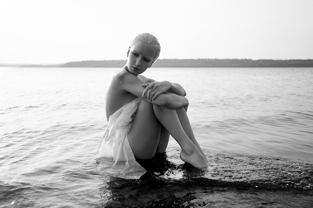 短い​散髪​の​アートヌードセクシー​な​ブロンド​は​、​日没時​に​湖​の​海岸​の​ビーチ​で​水​に​座っています​。​ウェットハ