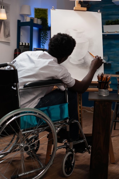 Art student in wheelchair sketching vase ilustration working at shadow using professional graphic tools during creative class. Young man illustrator drawing on canvas having inspiration in studio