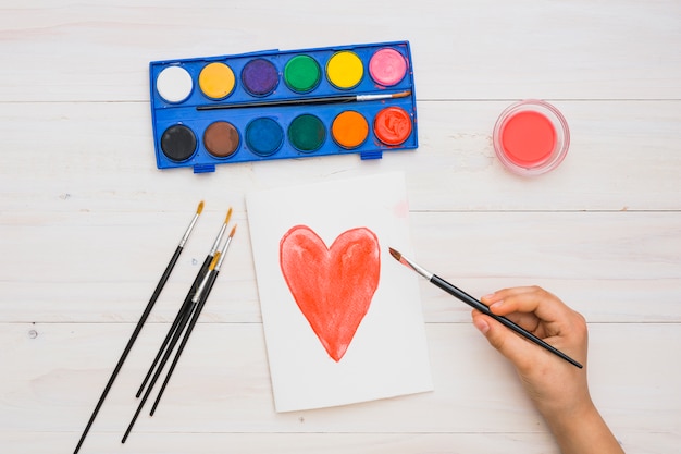 Free photo artist's hand holding paint brush on hand drawn heart shape painting over wooden surface