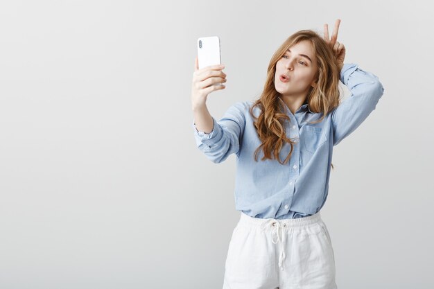 Not ashamed to be funny in front of camera. Good-looking positive feminine girl with blond hair in blue blouse, taking selfie while making faces and showing v sign behind head, aping over gray wall