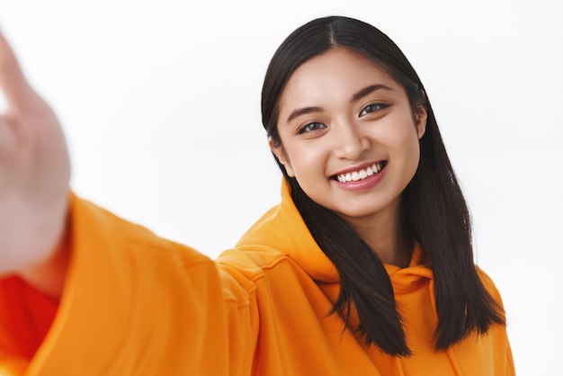 Asia beauty and fashion concept Closeup portrait beautiful asian girl in orange hoodie taking selfie holding camera with extended hand and smiling as make photo use new kawaii filters smiling