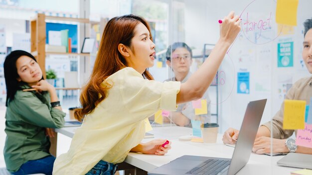 Asia businesspeople discussing business brainstorm meeting together share data and write on acrylic partition stand back in new normal office. Lifestyle social distancing and work after coronavirus.