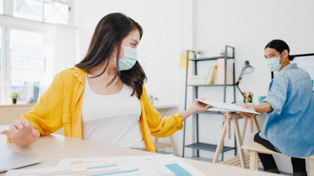 アジアのビジネスマンは、ウイルスの予防とオフィスでの距離を保ちながら文書を渡すための新しい通常の状況での社会的距離を保つためにフェイスマスクを着用します。コロナウイルス後のライフスタイルと仕事。