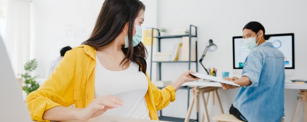 Asia businesspeople wear face mask for social distancing in new normal situation for virus prevention and passing documents with keeping a distance in office. Lifestyle and work after corona virus.