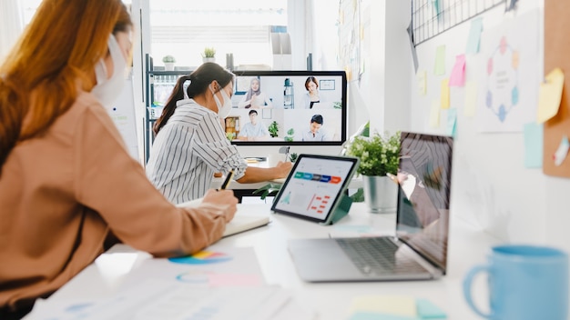 Asia businesspeople wear face mask using desktop talk to colleagues discussing brainstorm about plan in video call meeting in new normal office.