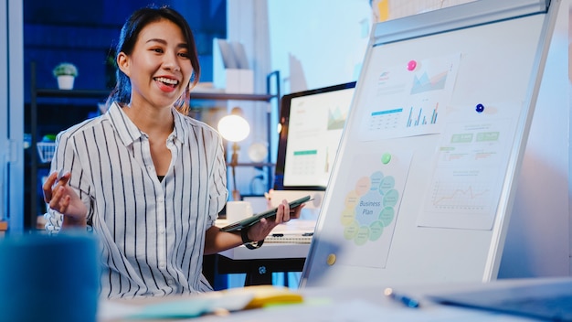 Asia businesswoman social distancing in new normal for virus prevention looking at camera presentation to colleague about plan in video call while work in office night.