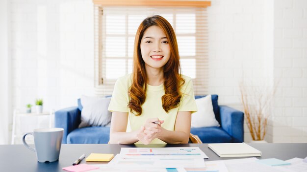 Asia businesswoman using computer laptop talk to colleagues about plan in video call meeting while working from home at living room. Self-isolation, social distancing, quarantine for corona virus.