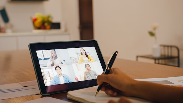 デジタルタブレットを使用しているアジアの実業家は、自宅のキッチンでリモートで作業しながら、ビデオ通話のブレインストーミングオンライン会議で計画について同僚と話します。