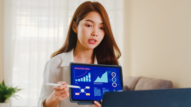 Free Photo asia businesswoman using laptop and tablet presentation to colleagues about plan in video call while working from home at living room. self isolation, social distancing, quarantine for coronavirus.
