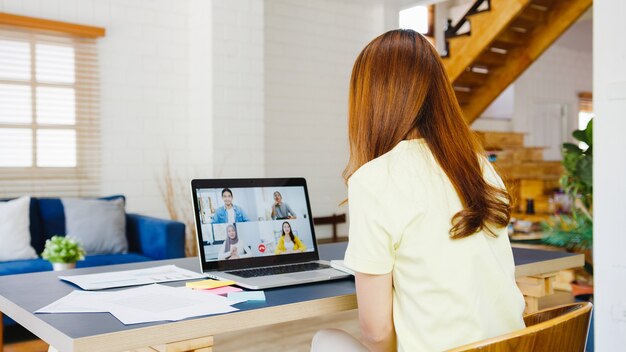 Asia businesswoman using laptop talk to colleagues about plan in video call meeting while working from home at living room. Self-isolation, social distancing, quarantine for corona virus prevention.