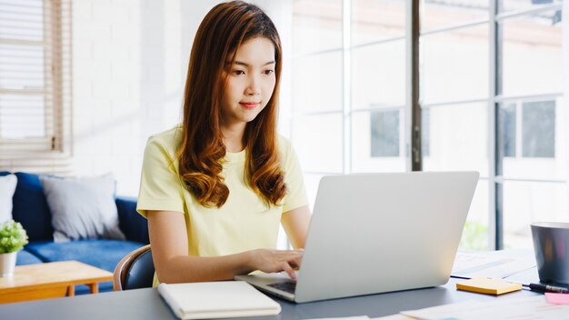 Asia businesswoman using laptop talk to colleagues about plan in video call while smart working from home at living room. Self-isolation, social distancing, quarantine for corona virus prevention.