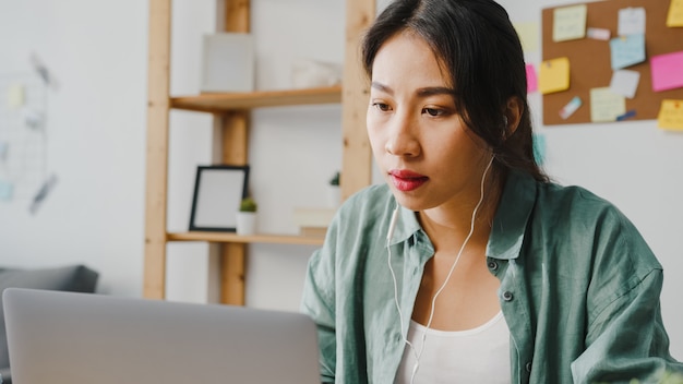 La donna d'affari asiatica che utilizza il laptop parla con i colleghi del piano in videochiamata mentre lavora in modo intelligente da casa in soggiorno