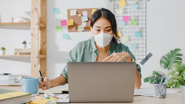 Free photo asia businesswoman wearing medical face mask using laptop talk to colleagues about plan in video call while working from home at living room.