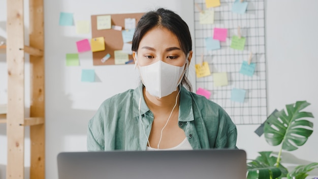 Азиатская деловая женщина в медицинской маске с ноутбуком разговаривает с коллегами о плане в видеозвонке, работая дома в гостиной.