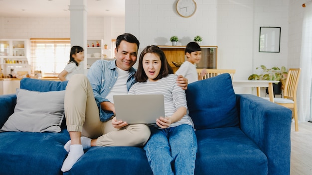 Asia family dad and mom sit on couch enjoy shopping online on laptop while daughter and son have fun shouting run around sofa in living room at home.