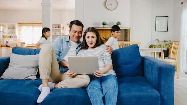 アジアの家族のお父さんとお母さんはソファに座ってノートパソコンでオンラインショッピングを楽しんでいますが、娘と息子は自宅のリビングルームでソファの周りを走り回って楽しんでいます。
