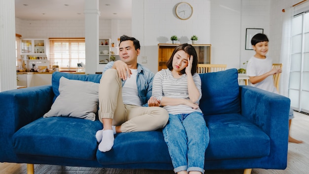 Asia family dad and mom sit on couch feels annoyed exhausted while daughter and son have fun shouting run around sofa in living room at home.