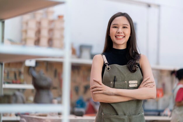 그녀의 워크샵 점토 조각 스튜디오에서 앞치마 손으로 자신감 있는 가슴을 가진 아시아 여성 도예가는 긍정적인 미소를 지으며 따뜻한 환영을 받으며 새로운 작업 컬렉션으로 새로운 공장 세라믹 워크샵을 시작할 준비가 되었습니다.