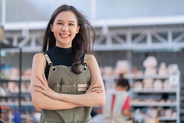 Asia donna ceramista con grembiule mano fiducioso petto nel suo laboratorio studio di scultura in argilla con positivo sorridente caloroso benvenuto pronto per iniziare un nuovo laboratorio di ceramica di fabbrica con una nuova collezione di lavori