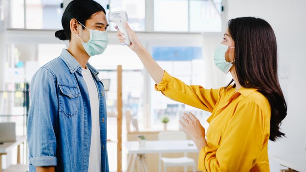 착용 보호 안면 마스크를 실시하는 아시아 여성 접수 원은 사무실에 들어가기 전에 고객의 이마에 적외선 온도계 검사기 또는 온도 건을 사용합니다. 코로나 바이러스 이후 생활 방식이 새로운 정상입니다.