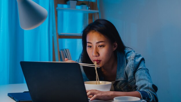 Asia freelance smart business women eating instant noodles while working on laptop in living room at home at night. Happy young Asian girl sitting on desk work overtime, enjoy relax time.