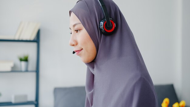 아시아 무슬림 여성 착용 헤드폰 시계 웨비나 온라인 과정을 듣고 집에서 화상 통화로 의사 소통합니다.