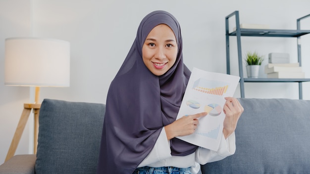 Foto gratuita la signora musulmana asiatica indossa l'hijab usa il computer portatile parla con i colleghi del rapporto di vendita nella riunione di videochiamata mentre lavora in remoto da casa in soggiorno. distanziamento sociale, quarantena per il virus corona.