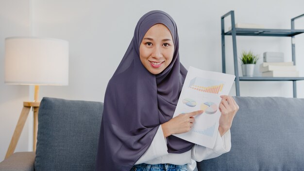 Азиатская мусульманка в хиджабе использует компьютерный ноутбук, разговаривает с коллегами об отчете о продажах на видеовстрече, когда удаленно работает из дома в гостиной. Социальное дистанцирование, карантин на вирус короны.