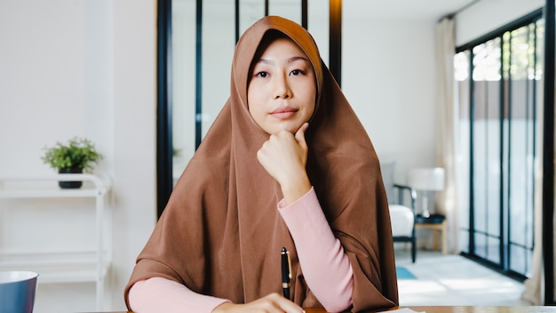 컴퓨터 랩톱을 사용하는 아시아 무슬림 여성은 거실에서 원격으로 작업하는 동안 화상 통화 회의 계획에 대해 동료에게 이야기합니다.