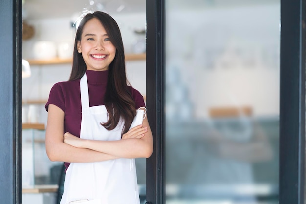 アジアの魅力的な女性カフェスタッフは、店先の入り口で封鎖が終わった後、前向きなサービスマインドで自信と幸せを持ってカフェレストランへの陽気な歓迎を笑顔で制服のエプロンを着ています