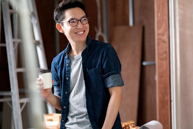 집 부지 개조 공사에서 일하는 아시아의 매력적인 안경 인테리어 디자이너는 청사진과 그리기 도구를 사용하여 책상에 미소와 자신감을 가지고 진행합니다.