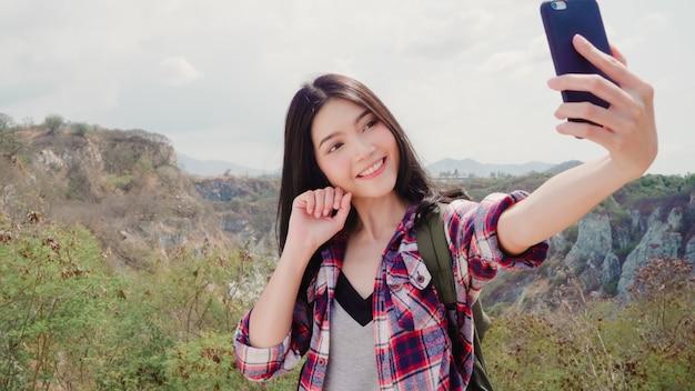 Asian backpacker woman selfie on top of mountain, young female happy using mobile phone taking selfie enjoy holidays on hiking adventure. 