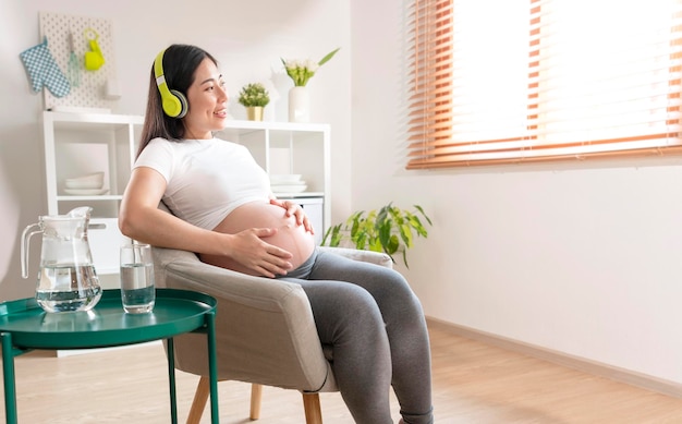 アジアの美しい妊娠中の女性の手が腹に愛撫し、自宅のソファに座ってヘッドフォンで音楽を聴いている妊娠出産の準備と期待の概念